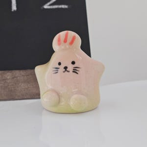 May include: A ceramic star-shaped figurine with a cartoon face. The figurine is primarily pink with a yellow-green base. It has black whiskers and a small black mouth. The top of the figurine has red stripes.