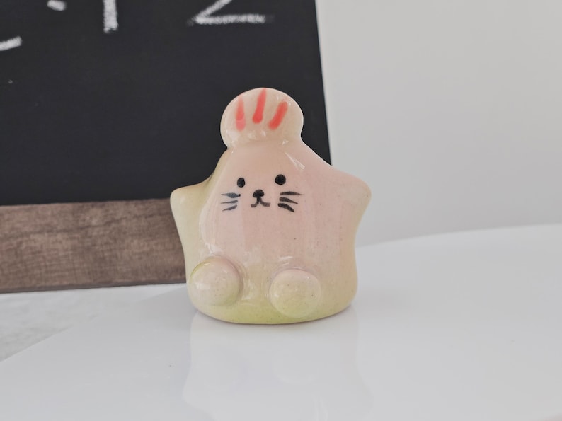 May include: A ceramic star-shaped figurine with a cartoon face. The figurine is primarily pink with a yellow-green base. It has black whiskers and a small black mouth. The top of the figurine has red stripes.