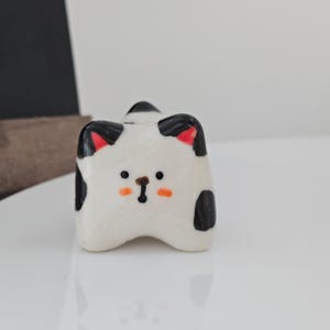 May include: A ceramic cat figurine with a white base, black spots, and red inner ears. The cat has an orange nose and black whiskers. The figurine is on a white surface.