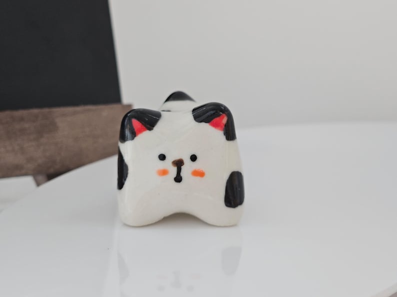 May include: A ceramic cat figurine with a white base, black spots, and red inner ears. The cat has an orange nose and black whiskers. The figurine is on a white surface.