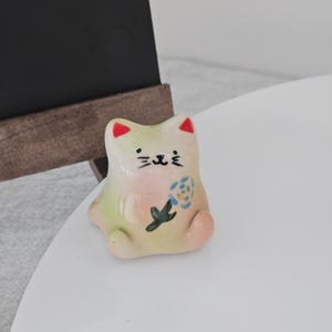 May include: A small, ceramic cat figurine with a whimsical design. The cat is primarily off-white with pink, green, and red accents. It features a painted face and a floral motif on its body. The figurine is sitting on a white surface.