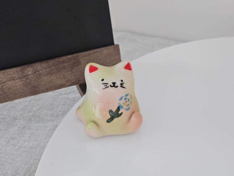 May include: A small, ceramic cat figurine with a whimsical design. The cat is primarily off-white with pink, green, and red accents. It features a painted face and a floral motif on its body. The figurine is sitting on a white surface.