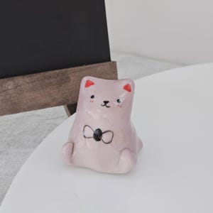 May include: A light pink ceramic cat figurine with a black bow tie and red accents on the ears. The cat is sitting with its legs tucked in. The figurine is on a white surface.