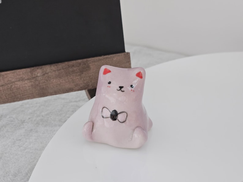 May include: A light pink ceramic cat figurine with a black bow tie and red accents on the ears. The cat is sitting with its legs tucked in. The figurine is on a white surface.