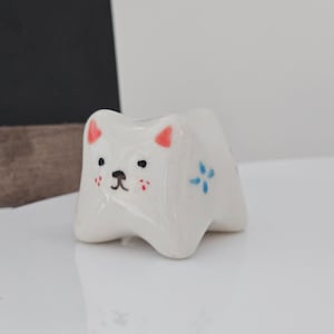 May include: A small, white ceramic cat figurine with a glossy finish. The cat has red ears, black eyes, and a black nose. It has small red cheeks and blue flower accents. The figurine is on a white surface.