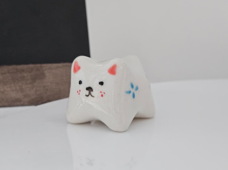 May include: A small, white ceramic cat figurine with a glossy finish. The cat has red ears, black eyes, and a black nose. It has small red cheeks and blue flower accents. The figurine is on a white surface.