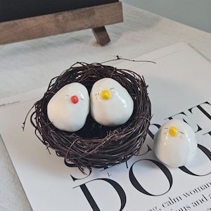 Peut inclure: Trois figurines d'oiseaux en céramique blanche aux yeux fermés et aux petits becs. Deux oiseaux sont nichés dans un nid de brindilles marron, tandis que le troisième est à côté du nid. Les oiseaux ont des accents rouges et jaunes. Le mot "DOOR" est visible.