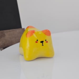 May include: A yellow ceramic cat figurine with orange ear accents and a black facial expression. The figurine is glossy and sits on a white surface. The cat has a cute, cartoonish design.