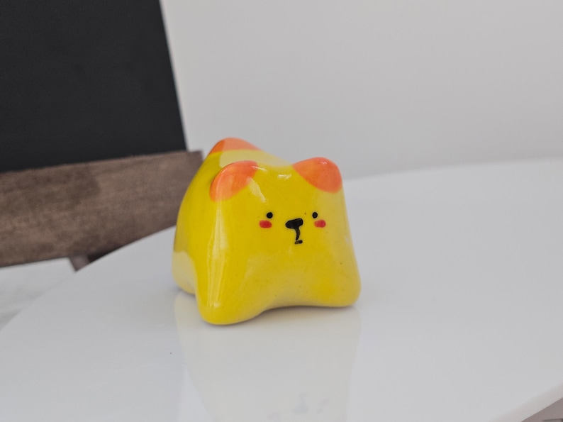 May include: A yellow ceramic cat figurine with orange ear accents and a black facial expression. The figurine is glossy and sits on a white surface. The cat has a cute, cartoonish design.