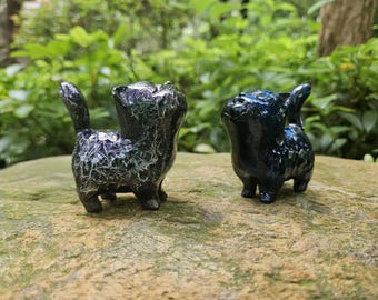 May include: Two black cat figurines with a speckled, iridescent finish. The cat on the left is looking up and has a tail. The cat on the right is looking to the left and has its ears perked up.