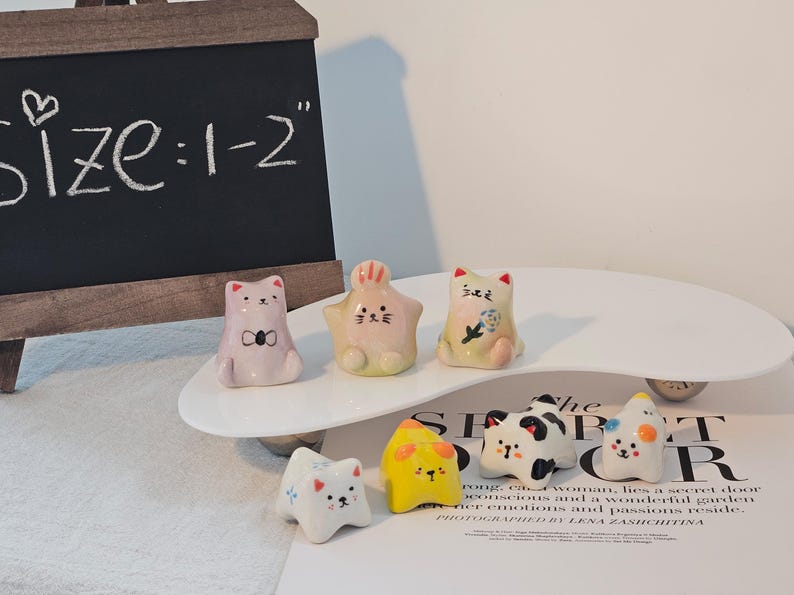 May include: A collection of small, ceramic figurines in the shape of cats and other animals. The figurines are in various colors, including pink, yellow, white, and black. The size is 1-2 inches. They are arranged on a white, curved platform.