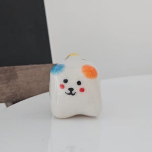 May include: A small, white ceramic dog figurine with a cartoonish design. The dog has black eyes, a black nose, and red cheeks. It has blue, orange, and yellow spots on its head. The figurine is on a white surface.