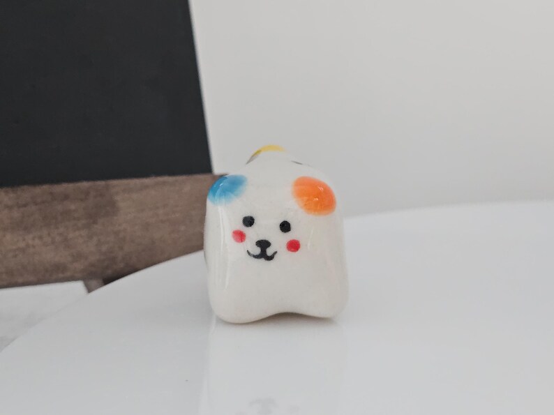 May include: A small, white ceramic dog figurine with a cartoonish design. The dog has black eyes, a black nose, and red cheeks. It has blue, orange, and yellow spots on its head. The figurine is on a white surface.