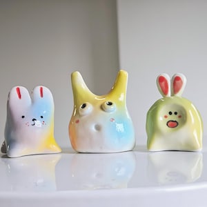 May include: Three whimsical ceramic figurines in pastel colors. The figurines have unique shapes, including a bunny-like figure with red-tipped ears, a yellow creature with large eyes, and a green figure with a surprised expression. They are displayed on a white surface.