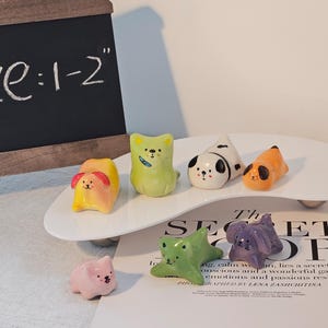 May include: A collection of colorful ceramic animal figurines, including dogs, cats, and a rabbit, displayed on a white, oval-shaped tray. The figurines are approximately 1-2 inches in size. A small chalkboard with the text "Size: 1-2" is in the background.