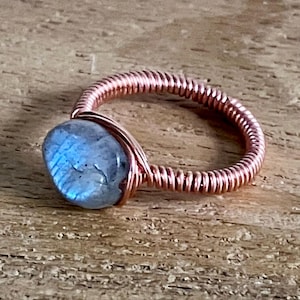 May include: A handmade copper wire ring with a blue gemstone. The ring's band is coiled, and the gemstone is oval-shaped. The ring is set on a wooden surface.