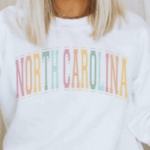 Puede incluir: Sudadera blanca de manga larga con el texto "NORTH CAROLINA" en colores arco&iacute;ris.