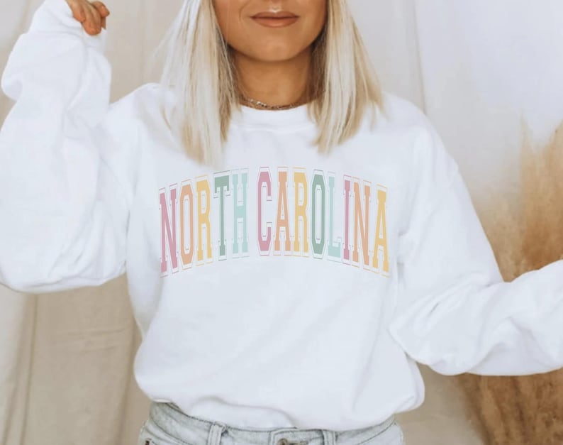 Puede incluir: Sudadera blanca de manga larga con el texto "NORTH CAROLINA" en colores arco&iacute;ris.