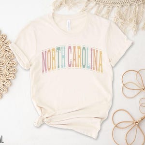 Puede incluir: Una camiseta de color natural con el texto "NORTH CAROLINA" en colores arco&iacute;ris.
