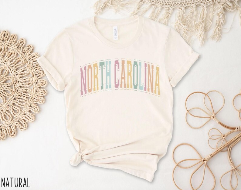 Puede incluir: Una camiseta de color natural con el texto "NORTH CAROLINA" en colores arco&iacute;ris.