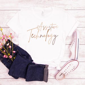 May include: White t-shirt with the text "Assistive Technology" written in a cursive font in brown and gold.