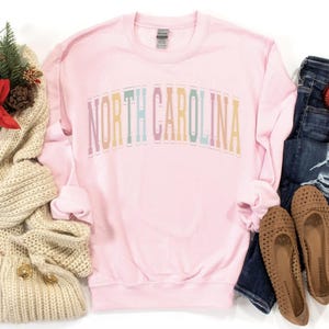 Puede incluir: Sudadera rosa de manga larga con el texto "NORTH CAROLINA" en colores arco&iacute;ris.