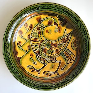 May include: A round ceramic plate featuring a central illustration of a bird in profile, rendered in shades of yellow, brown, and green. The bird is surrounded by stylised plant motifs, and the rim is decorated with a dark green repeating pattern.