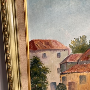 May include: Oil painting of a European village scene with buildings featuring red tile roofs and light-colored walls. The artwork is framed in a gold-colored ornate frame, with a neutral-colored fabric matting.