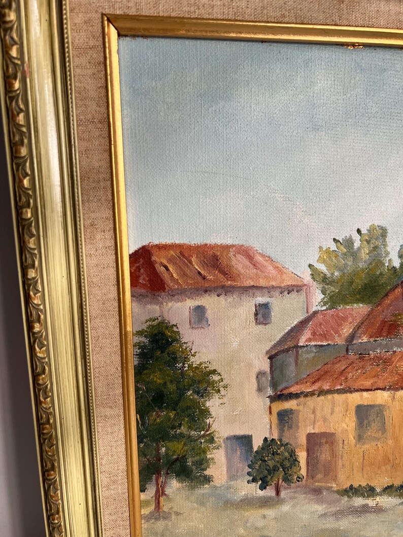 May include: Oil painting of a European village scene with buildings featuring red tile roofs and light-colored walls. The artwork is framed in a gold-colored ornate frame, with a neutral-colored fabric matting.