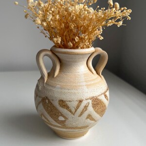 May include: A small, beige ceramic vase with two handles and a decorative brown pattern. The vase is filled with dried, light brown flowers. The vase has a rustic, handmade appearance.
