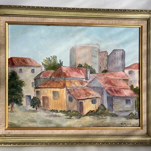 May include: Oil painting of a cityscape with buildings and red-tiled roofs, set against a light blue sky. The artwork is framed in a gold-colored ornate frame. Signed by M. Woodward.