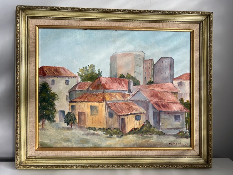 May include: Oil painting of a cityscape with buildings and red-tiled roofs, set against a light blue sky. The artwork is framed in a gold-colored ornate frame. Signed by M. Woodward.