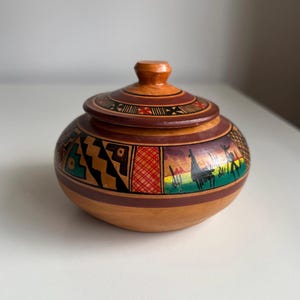 May include: A small, round, wooden jar with a lid. The jar is painted with colourful geometric and landscape designs in red, black, green, and yellow. The lid has a small knob on top.