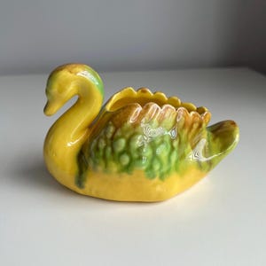 May include: A ceramic swan-shaped planter or trinket dish featuring a glossy finish. The body is primarily bright yellow with blended patches of green and brown along the textured wing area. The neck is curved, and the top has an open cavity.