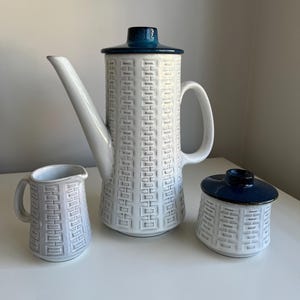 May include: A ceramic coffee set consisting of a tall coffee pot with a lid, a creamer, and a lidded sugar bowl. Each piece features a white body with a repeating embossed geometric pattern and deep blue glazed lids and accents.