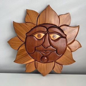 May include: A wooden sunburst wall hanging with a smiling face. The sun's face is a dark brown, with light brown eyes and a small, round nose. The petals are a lighter brown, creating a layered effect. The piece is approximately 30 cm in diameter.