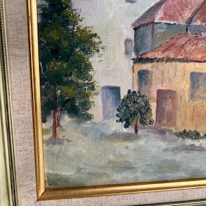 May include: Oil painting of a European village scene. The artwork features buildings with red tile roofs and light-colored walls. Trees with green foliage are in the foreground. The painting is in a gold-colored frame.