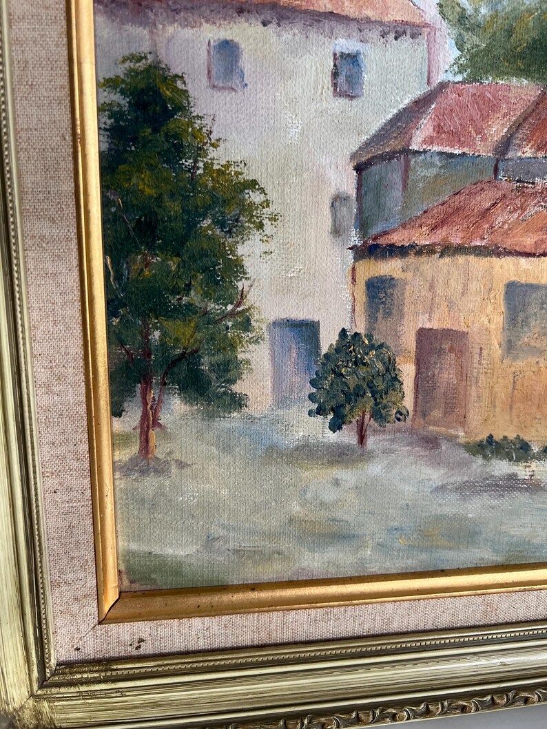 May include: Oil painting of a European village scene. The artwork features buildings with red tile roofs and light-colored walls. Trees with green foliage are in the foreground. The painting is in a gold-colored frame.