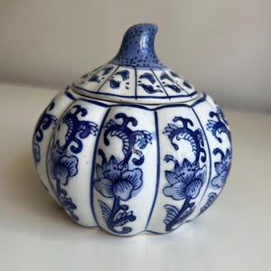 May include: A white ceramic lidded jar with a pumpkin-like ribbed shape, decorated with blue floral patterns in vertical panels. The lid features a blue speckled stem-like handle, and the body displays repeating blue botanical motifs.