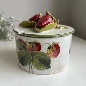 May include: A round ceramic jar with a white crackle glaze finish, featuring a painted strawberry and green leaf motif on the side. The lid is topped with a three-dimensional sculpted red strawberry and green leaves, including a small notch for a spoon.