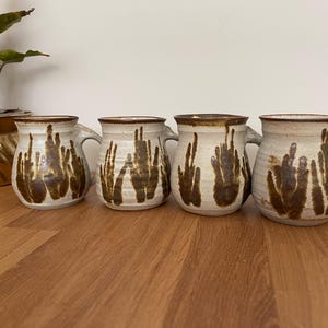 May include: Four brown and white ceramic mugs with a unique hand-painted design. The mugs have a rounded shape and a handle on the side.
