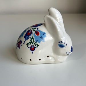 May include: A white ceramic bunny ornament decorated with blue and red floral patterns. The bunny has blue eyes and a dotted nose. Small holes are visible on the side, possibly for use as a salt or pepper shaker. A decorative item.
