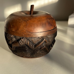 May include: A wooden apple-shaped box, featuring a carved design around the base and a stem. The box is crafted from dark brown wood, with a lid that fits securely. The carved design shows figures and foliage, adding detail to the piece.