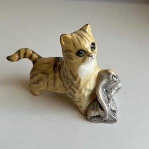 May include: A ceramic figurine of a tan and brown tabby cat with dark stripes, green eyes, and a white chest patch. The cat is standing on all four paws with its tail raised, holding a small, grey, textured object with its front paws.