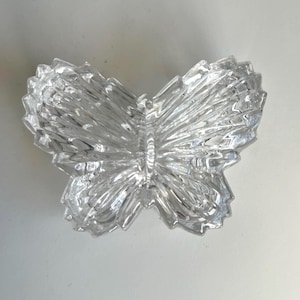 May include: A clear glass butterfly figurine featuring a textured, faceted surface that mimics the appearance of wings. The object is shaped like a butterfly with a central body and four distinct wing sections, all crafted from transparent glass.