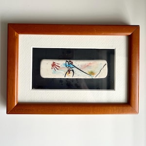 May include: A rectangular ceramic tile featuring a hand-painted abstract design with a red sun, a black stick figure, and colourful brushstrokes, mounted on a black background within a light brown wooden frame with a white mat border.