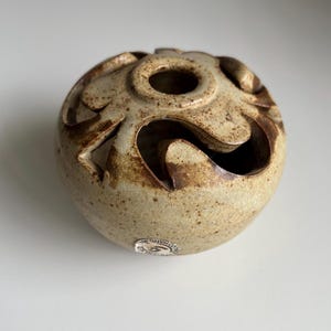 May include: A small, round, ceramic decorative object with a speckled, tan and brown finish. The top has a central hole and cut-out designs around the sides. A small sticker is visible on the side.