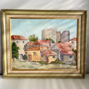 May include: Oil painting of a village scene with buildings and red roofs, set in a gold ornate frame. The artwork depicts a cluster of buildings with a few trees and a light blue sky. Signed by M. Woodward.