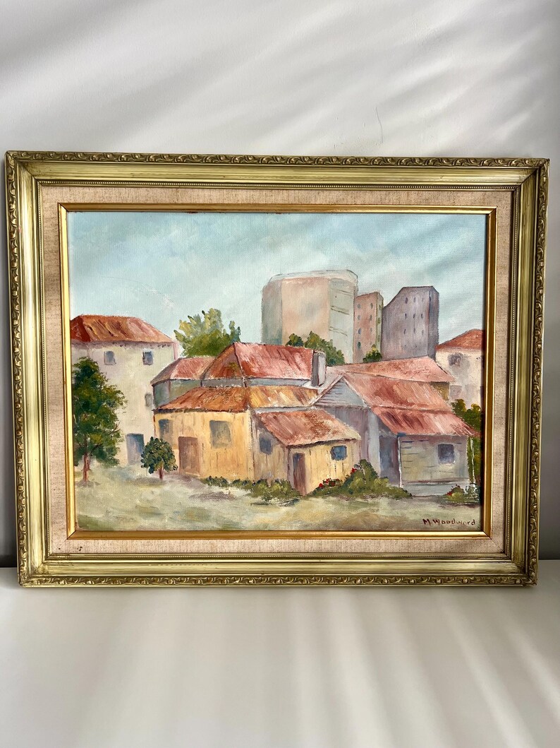 May include: Oil painting of a village scene with buildings and red roofs, set in a gold ornate frame. The artwork depicts a cluster of buildings with a few trees and a light blue sky. Signed by M. Woodward.