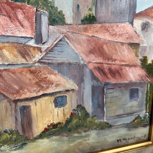 May include: Oil painting of a European village scene with red-tiled roofs and light-colored buildings. The artwork is framed in a gold-colored frame and signed by M. Woodward. The painting depicts a sunny day with a clear sky.
